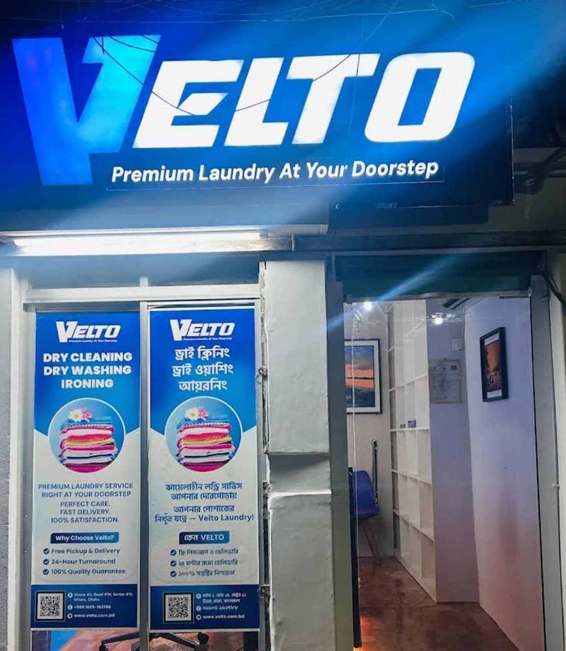 The Velto Sector 11 outlet at night: the lit Velto sign above the entrance, with service posters in the window.