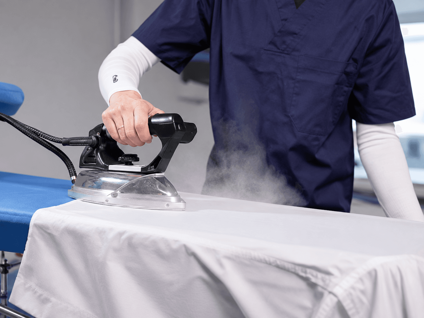 A person holding a stack of neatly folded shirts beside a steam iron on an ironing board.